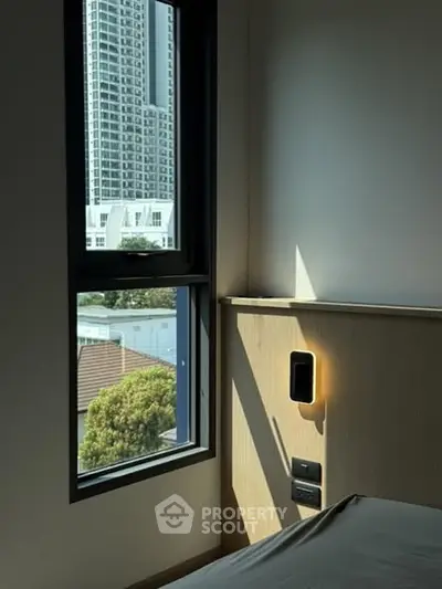 Modern bedroom with city view through large window, featuring sleek wall lighting.