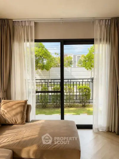Elegant living room with large window overlooking a lush garden, featuring cozy beige sofa and sheer curtains.