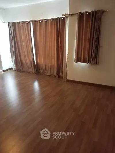 Spacious empty room with wooden flooring and large windows with curtains.
