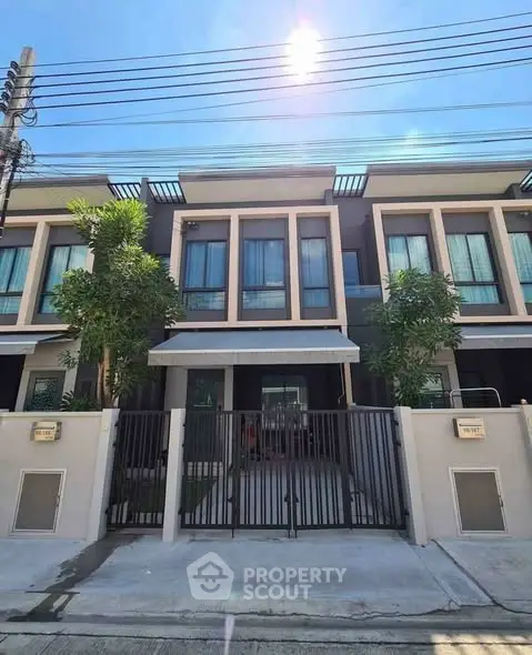 Modern townhouse exterior with gated entrance and lush greenery under a bright blue sky.
