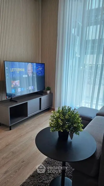 Modern living room with TV, stylish furniture, and large window with sheer curtains.