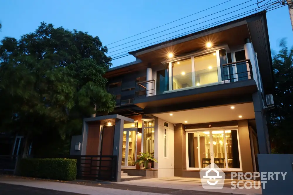 Modern two-story house with large windows and evening lighting, surrounded by lush greenery.