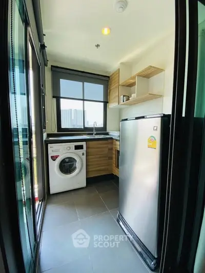 Modern compact kitchen with washing machine and fridge, featuring sleek cabinetry and large window view.