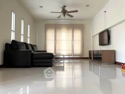 Spacious living room with sleek black sofa and mounted TV, featuring glossy tiled floors and ceiling fan.