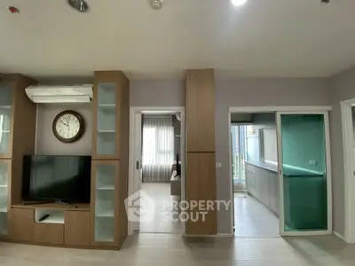 Modern apartment interior with TV, clock, and sliding doors leading to a bright kitchen.