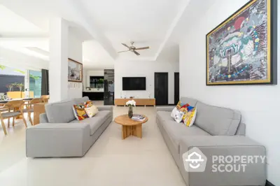 Spacious living room with modern grey sofas, pristine white floors, and an open layout leading to a dining area, accentuated by a vibrant wall art.