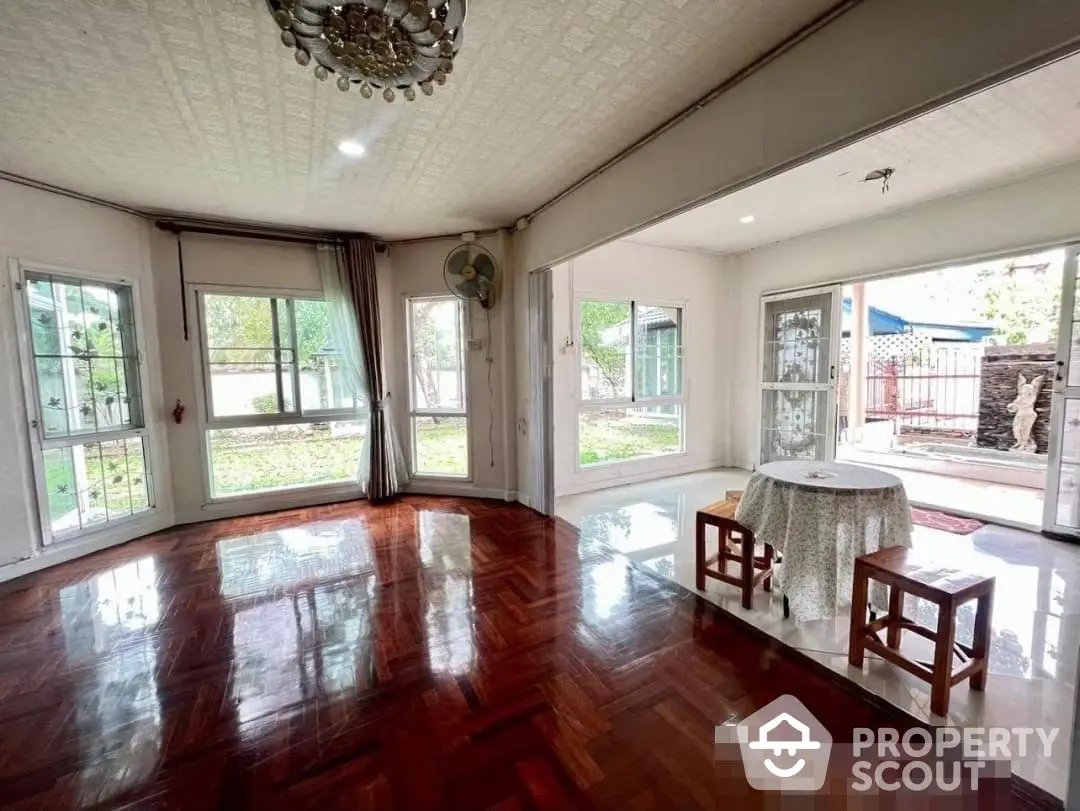 Spacious living room with gleaming hardwood floors, elegant chandelier, and abundant natural light from French doors leading to a lush garden.