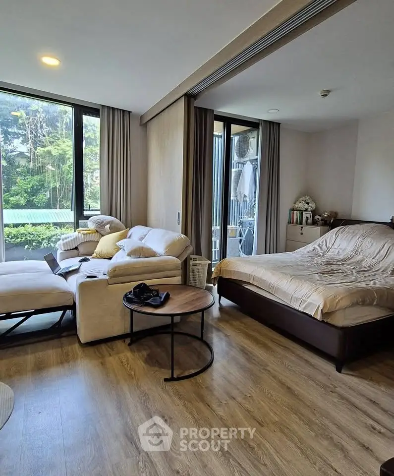 Spacious studio apartment with cozy living area and large windows offering lush green views.