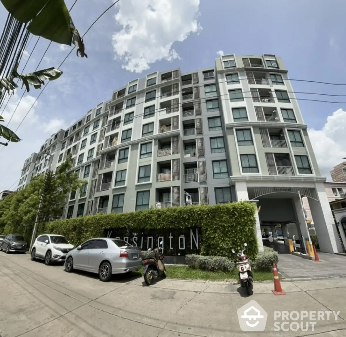 Modern multi-story residential building with lush greenery and parked vehicles in urban setting.