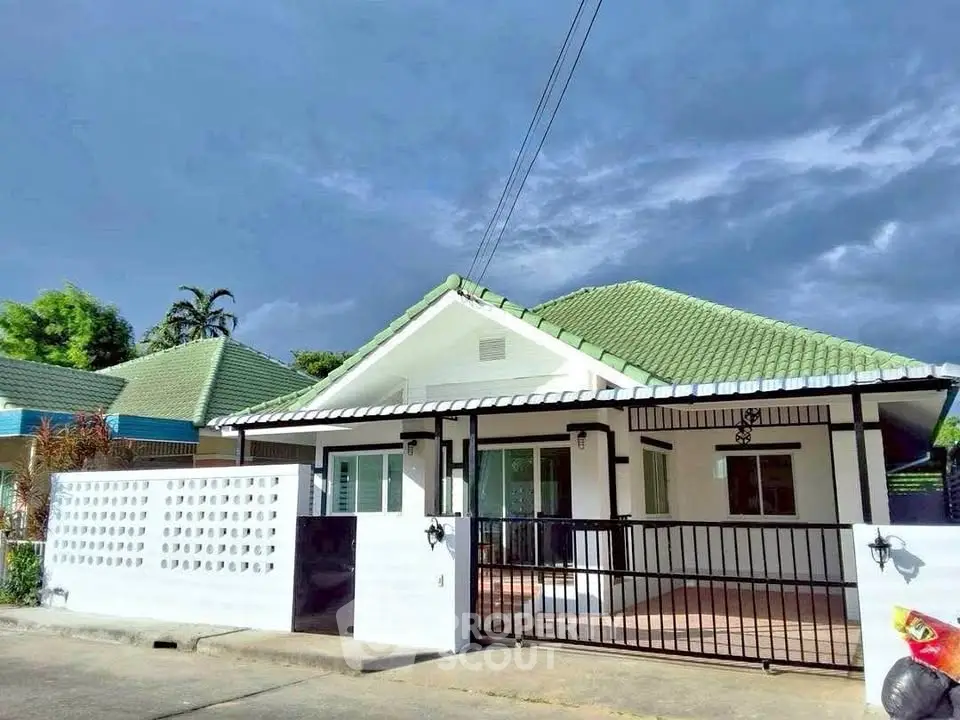 Charming single-story house with green roof and gated entrance under a dramatic sky.