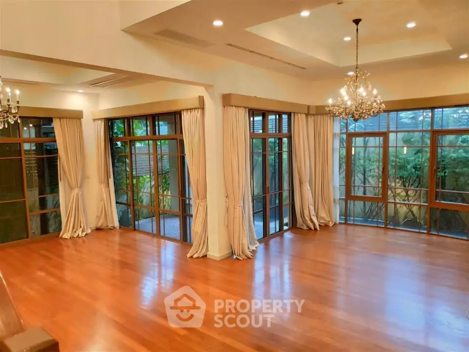 Spacious living room with elegant chandeliers and large windows overlooking lush greenery.