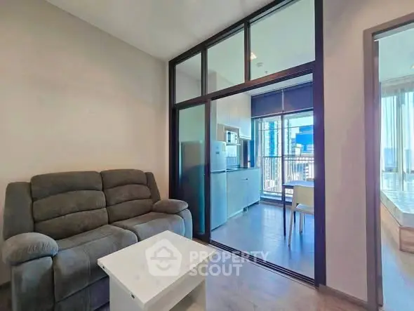 Modern living room with glass partition leading to a stylish kitchen and dining area, featuring sleek furniture.