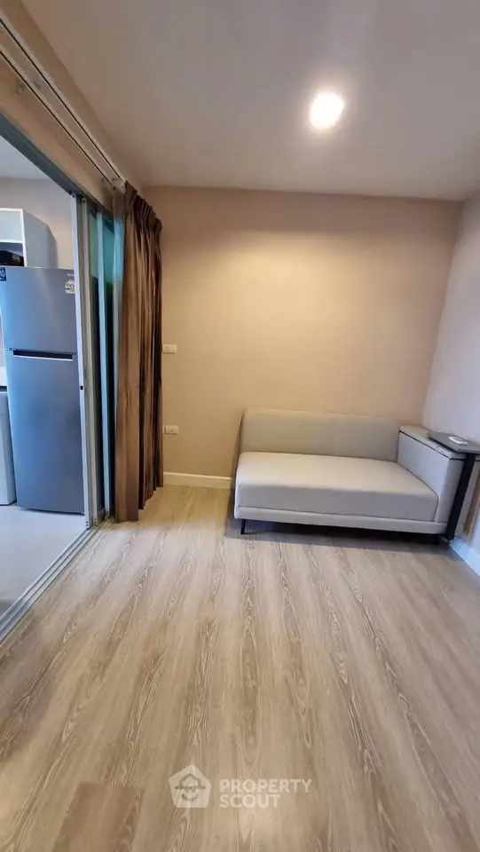 Modern living room with wooden flooring and compact sofa, adjacent to a kitchen with stainless steel fridge.