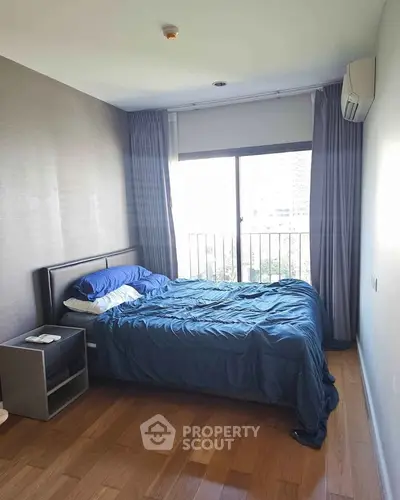 Cozy bedroom with wooden flooring and large window offering natural light and city view.