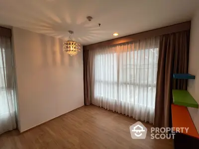 Spacious living room with elegant chandelier lighting, large windows draped with sheer and opaque curtains, and warm wooden flooring.