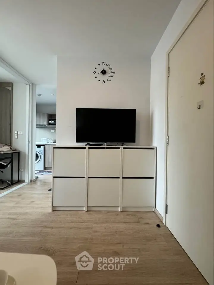 Modern living room with sleek TV stand and wall clock, open view to kitchen with washing machine.