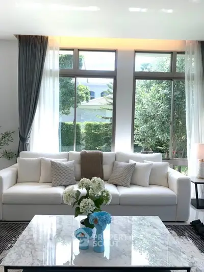Elegant living room with large windows, plush white sofa, and stylish decor, offering a serene garden view.