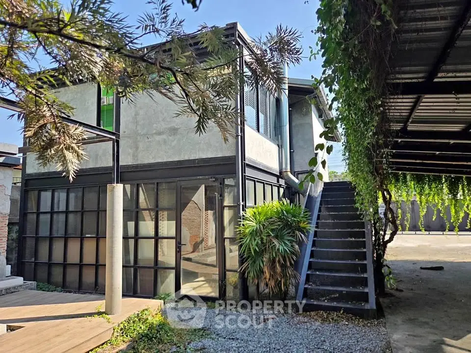 Modern industrial-style home with large windows and outdoor staircase, surrounded by lush greenery.