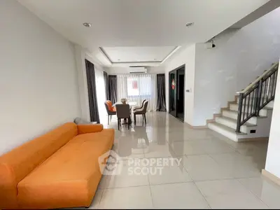 Spacious living room with modern orange sofa and dining area, featuring elegant staircase and large windows.
