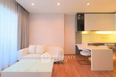Modern living room with open kitchen, featuring sleek white furniture and warm wooden flooring.