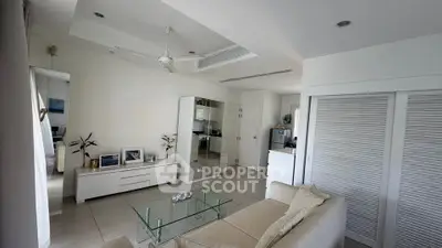 Modern living room with sleek white furniture and glass coffee table, open layout to kitchen area.