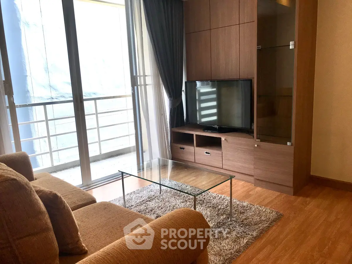 Modern living room with glass coffee table and large TV, featuring floor-to-ceiling windows and wooden flooring.