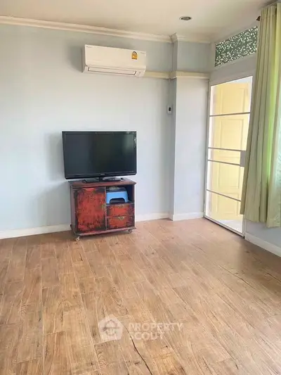 Spacious living room with wooden flooring and natural light, featuring a TV and air conditioning.