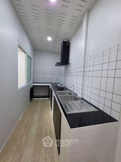 Modern kitchen with sleek black countertops and gas stove in a narrow layout, featuring white tiled walls and wooden flooring.