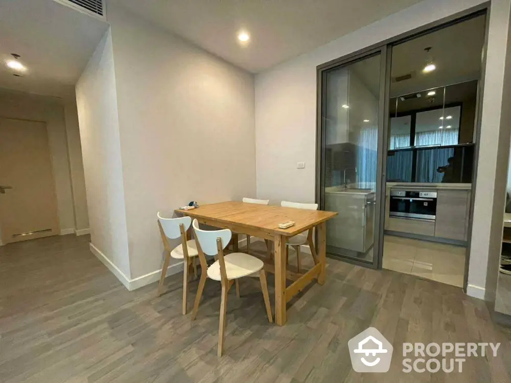 Modern dining area with wooden table and chairs next to a sleek kitchen with glass partition.