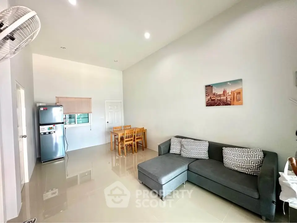 Modern living room with sleek furniture and open dining area, featuring a stylish gray sofa and stainless steel fridge.