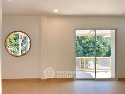 Bright living room with circular window and sliding glass door leading to a balcony with lush greenery view.