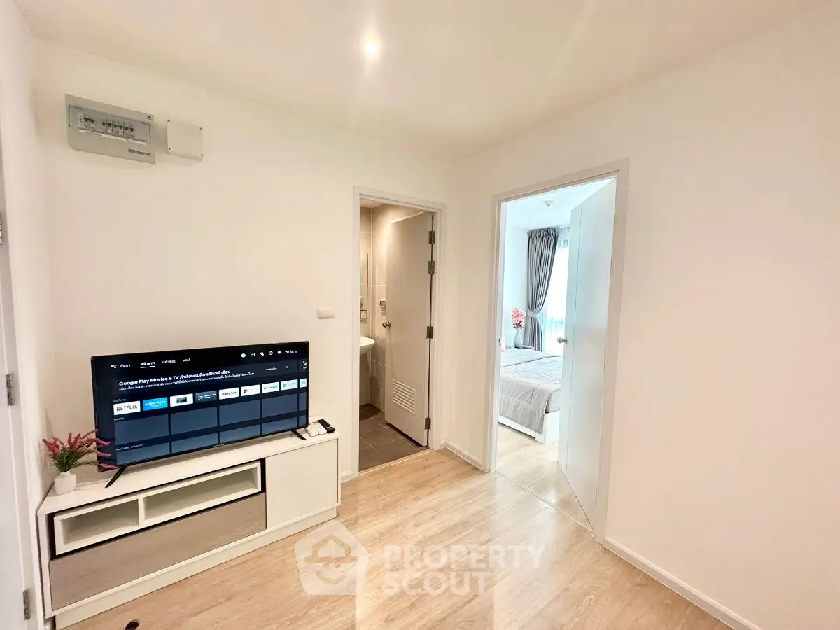 Modern apartment interior with TV, light wood flooring, and view into a cozy bedroom.