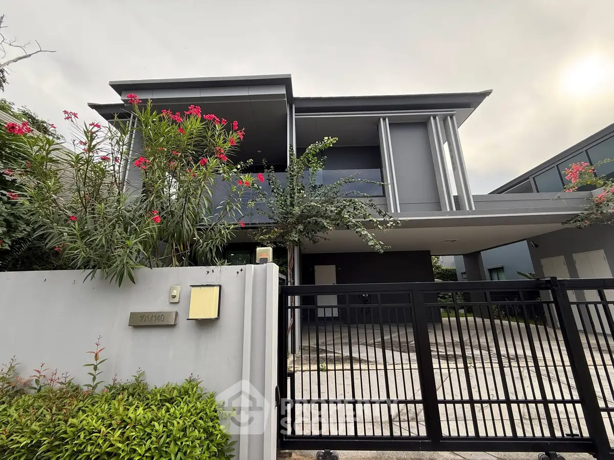 Modern two-story house with sleek design and gated entrance, surrounded by lush greenery.
