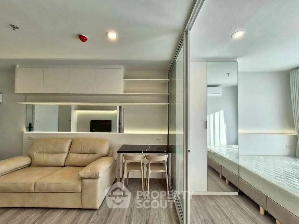 Modern living room with beige sofa and glass partition leading to bedroom, featuring sleek design and ample lighting.