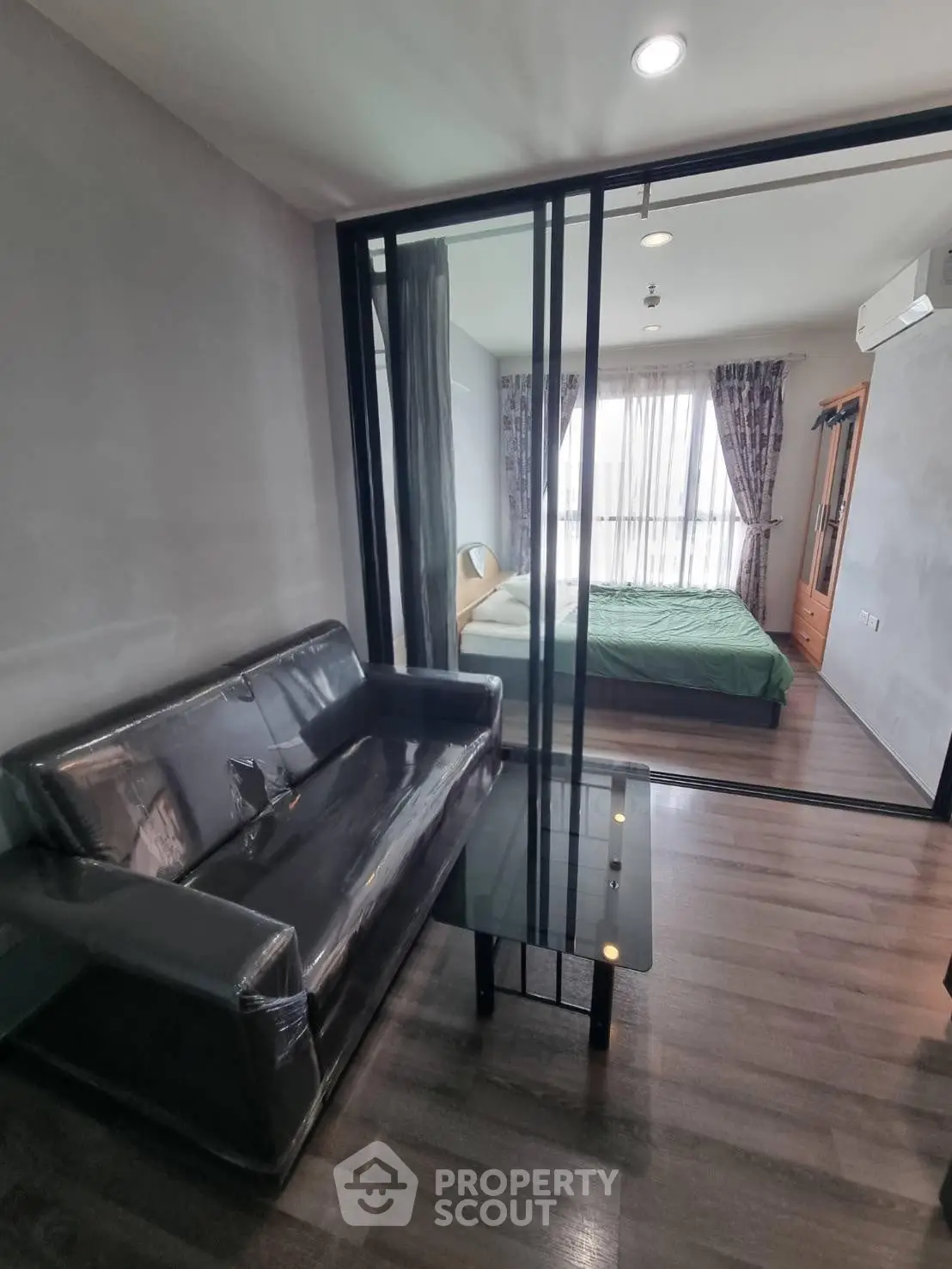 Modern apartment with glass partition separating living room and bedroom, featuring sleek furniture and natural light.