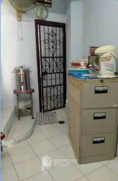 Compact kitchen area with metal filing cabinet and water dispenser, featuring tiled flooring and a secure door.
