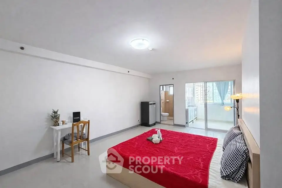 Spacious bedroom with modern decor and natural light, featuring a cozy red bedspread and a small study area.