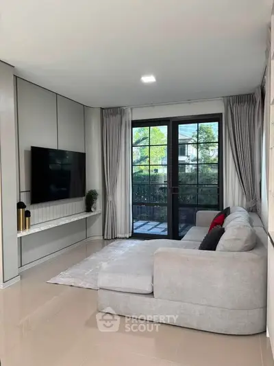 Modern living room with large sofa, wall-mounted TV, and glass doors leading to a balcony with greenery view.