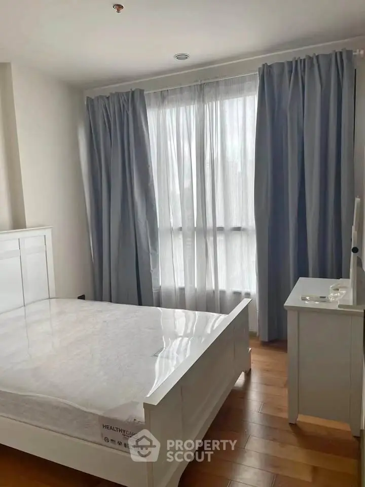 Bright bedroom with large window and elegant curtains, featuring a white bed and wooden flooring.