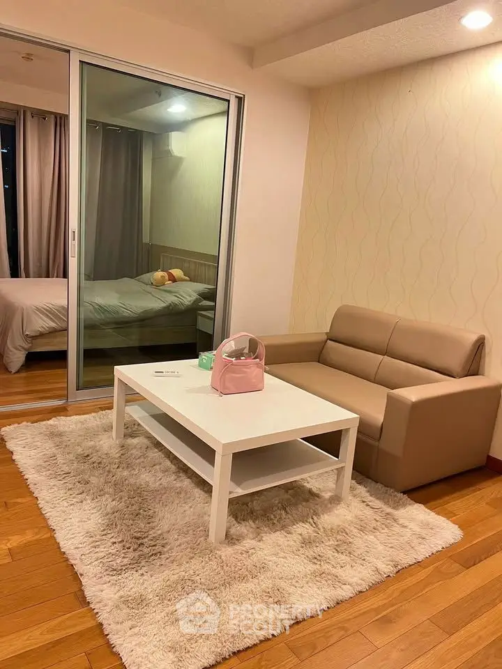 Cozy living room with beige sofa, white coffee table, and view into a modern bedroom through sliding glass doors.