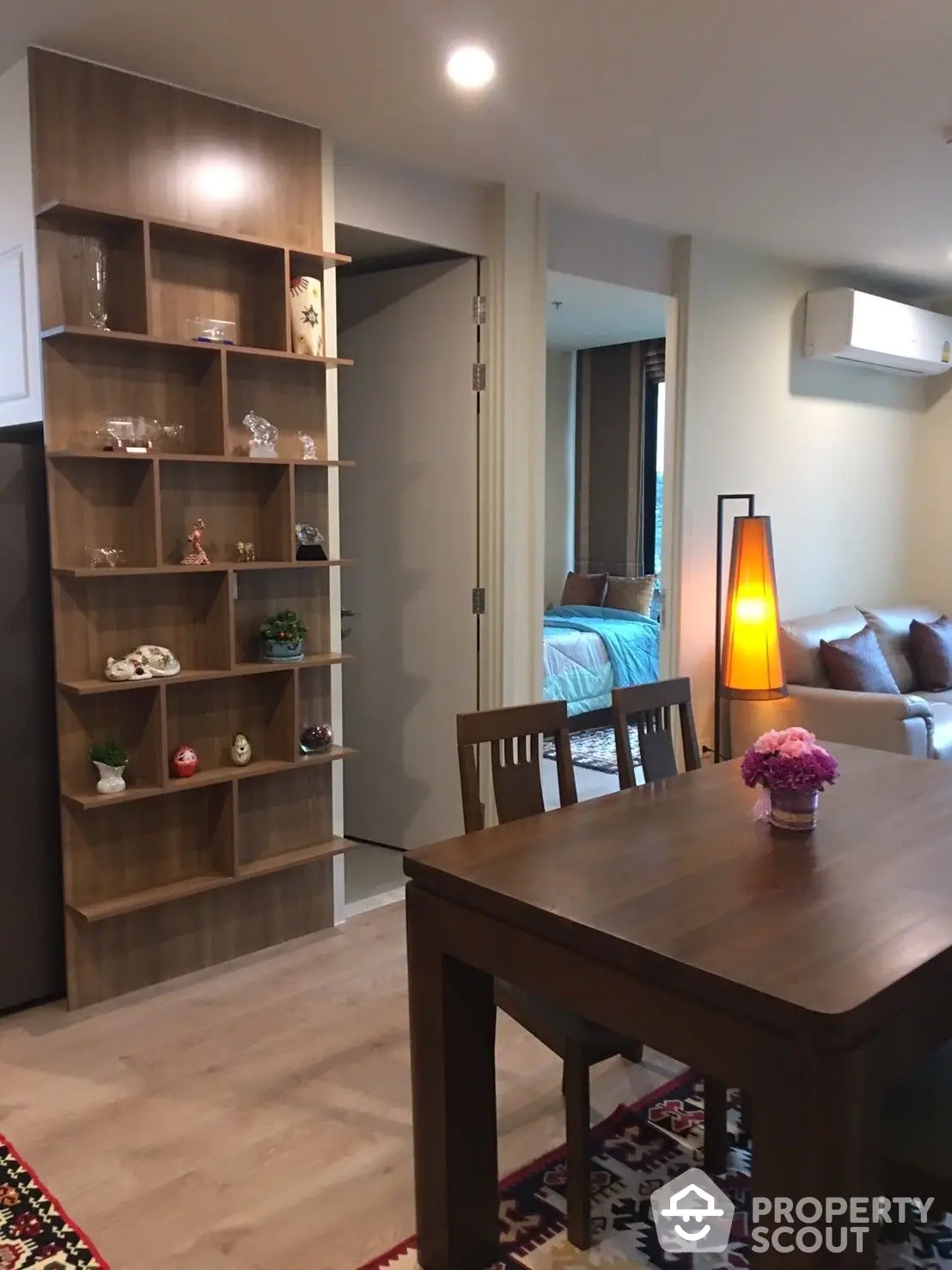 Cozy apartment interior with warm lighting, showcasing a wooden dining table, shelving unit, and a glimpse into the comfortable bedroom.