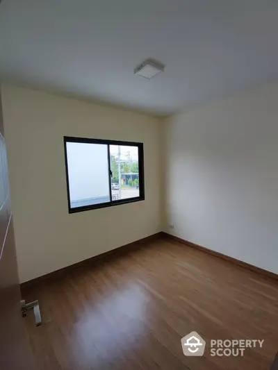 Spacious and well-lit empty room with polished wooden flooring and a large window offering a street view, ideal for a personalized living space.