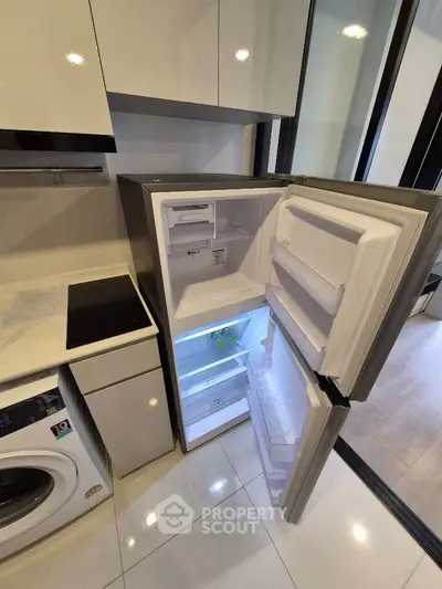 Modern kitchen with open fridge and washing machine, sleek cabinetry and glossy tiles.