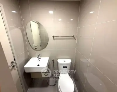 Modern bathroom with sleek fixtures and neutral tiles, featuring a stylish oval mirror and compact sink.