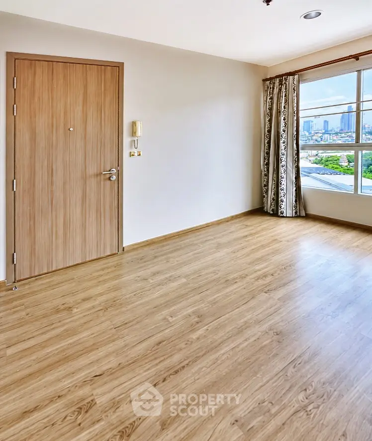 Spacious empty room with wooden flooring and large window offering city views.