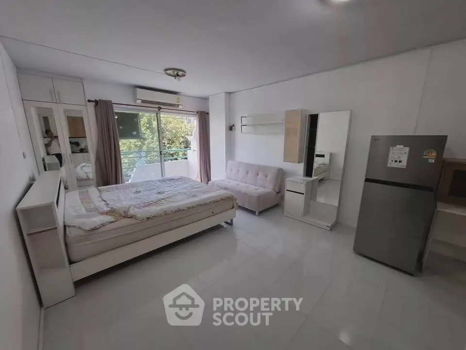 Spacious studio apartment with modern furnishings and balcony view, featuring a cozy bed and sleek refrigerator.