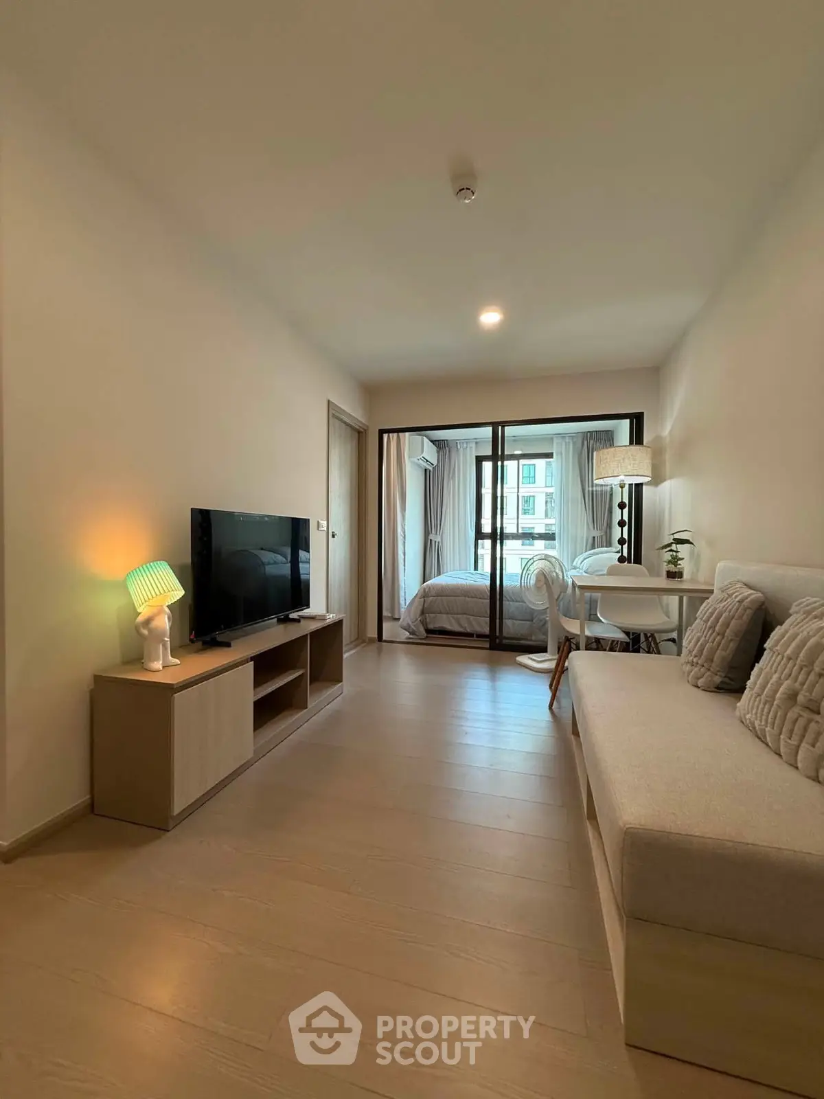 Modern living room with cozy seating and TV, leading to a bright bedroom with large windows.