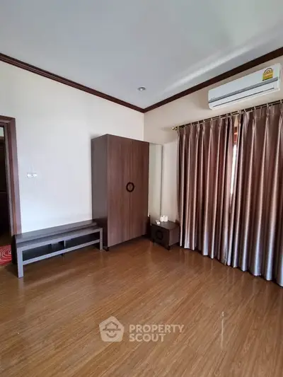Spacious bedroom with wooden flooring and modern furniture, featuring a large window with elegant curtains.