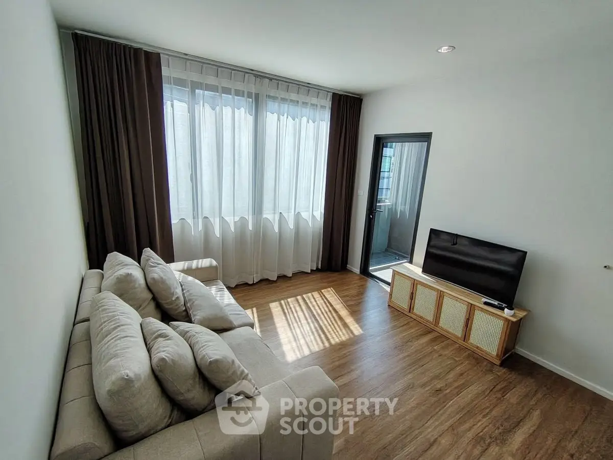 Bright living room with modern sofa and TV, featuring large window and wooden flooring.
