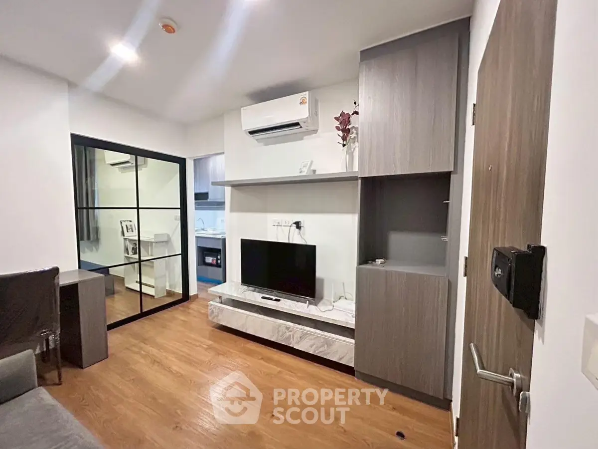 Modern living room with sleek wooden flooring, built-in TV unit, and air conditioning, leading to a compact kitchen.